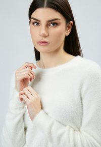 Maglione bianco e morbido con scollo rotondo e maniche lunghe. Il tessuto sembra morbido e testurizzato, con una finitura leggermente pelosa.