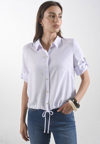 Lichtpaarse blouse met korte mouwen, kraag, knopen aan de voorkant, trekkoordzoom en opgerolde manchetten, gecombineerd met donkerblauwe jeans en armbanden.
