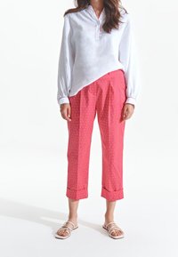 Kvinna iklädd en vit långärmad blus, rosa mönstrade cropped byxor med manschetter och vita flat sandals med remmar, står framför en vit bakgrund.