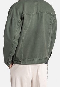 Groene spijkerjasje met een ontspannen pasvorm, voorzien van een gerafelde onderzoom met knopen en zichtbare stiksels. Gladde textuur, zonder zichtbare patronen.