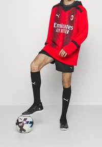 Rotes Fußballtrikot mit langen Ärmeln, schwarzen Akzenten und Logo, kombiniert mit schwarzen Shorts, Socken und Schuhen, steht auf einem gemusterten Fußball.