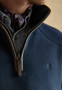 Detailný záber na osobu, ktorá má na sebe tmavomodrý fleece pulóver s hnedým zipsom a pod ním pruhované tričko, ktoré je doplnené kvetinovou šatkou okolo krku.