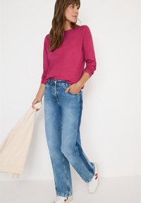 Fuchsia långärmad topp med avslappnad passform, i kombination med ljusblå rak ben-jeans. Vita sneakers och en ljuscremefärgad tygprodukt i handen.