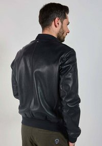 Veste aviateur en cuir noir au design épuré, avec des poignets et un ourlet côtelés, et une finition texturée. Présente un petit logo sur le bras gauche.