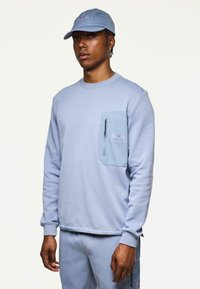 Joven vestido con sudadera azul clara, pantalones a juego y una gorra, todos con bolsillos con cremallera y la marca "Weekend Offender".