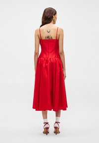 Κόκκινο αμάνικο φόρεμα με εφαρμοστό bodice και φουσκωτή φούστα ως το γόνατο. Λείο ύφασμα, φερμουάρ στην πλάτη, συμπληρωμένο από μεσαίες γόβες.