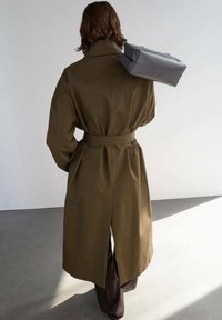Trench coat verde oliva con vita ampia, caratterizzato da una fessura sul retro e spalle scese, abbinato a pantaloni marrone scuro e una borsa grigia.