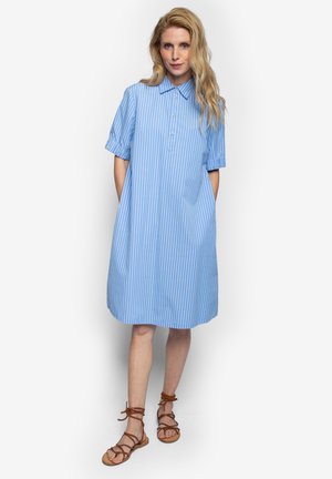 Blondynka stoi z rękami w kieszeniach, ubrana w niebiesko-białą sukienkę koszulową do kolan w pionowe paski oraz brązowe płaskie sandały na sznurowadła.