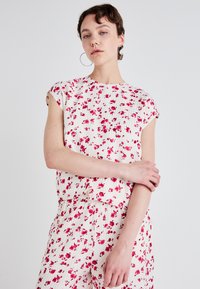Blusa floreale a maniche corte bianca con motivo di fiori rossi. Tessuto leggero con una finitura texturizzata e leggermente plissettata. Collo rotondo.