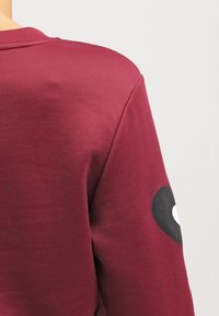 Bordowy sweatshirt z prążkowanym dekoltem i czarnym okrągłym wzorem na rękawie. Wykonany z gładkiego, lekkiego materiału.