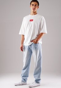 Weißes, übergroßes T-Shirt mit rotem Etikett, kombiniert mit hellblauen Jeans. Das Outfit umfasst weiße Sneaker mit roten Akzenten.