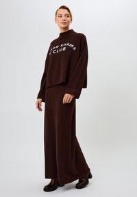 Ruskoharma neulepaita, jossa on "Hyvän karmaklubi" -teksti, yhdistettynä sopivaan ruskeaan maksimekkoon. Toinen kaulus ja leveät hihat.