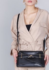 Sort tekstureret crossbody-taske med gyldne detaljer og et zigzag-mønster, der har en lang justerbar rem og en elegant, kompakt form.