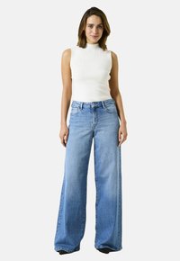 Een vrouw staat tegen een witte achtergrond en draagt een mouwloos wit geribd topje en lichtblauwe wijde jeans.