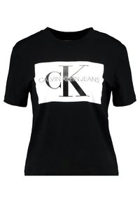 Schwarzes Baumwoll-T-Shirt mit einem weißen rechteckigen Panel, auf dem "CALVIN KLEIN JEANS" und das stilisierte "ck"-Logo in Silber zu sehen sind. Kurze Ärmel, Rundhalsausschnitt.