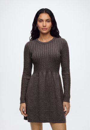 Femme portant une robe en tricot ajustée à manches longues, gris foncé, avec des motifs torsadés sur la partie supérieure et une jupe unie.