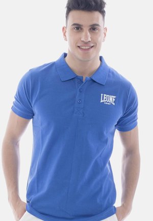 Jonge man glimlachend, draagt een blauw poloshirt met het logo "LEONE 1947", staat met handen in de zakken tegen een witte achtergrond.