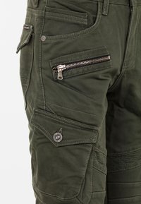 Grüne Cargo-Hosen mit mehreren Taschen, einschließlich einer Reißverschluss-Tasche, und strukturierten Nähten auf dem Stoff für zusätzlichen Detailreichtum.
