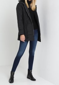 Donna con una giacca trapuntata nera, top nero, jeans skinny blu e stivaletti neri in piedi contro uno sfondo bianco.