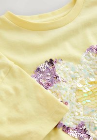 Tissu jaune avec un motif d'étoile pailletée en nuances de blanc et violet sur un vêtement à manches longues, montrant l'encolure et l'ourlet de la manche.