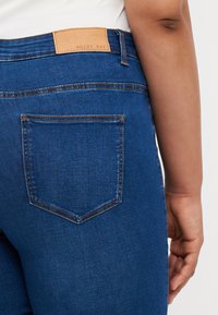 Blue denim jeans with a leather patch labeled "NOISY MAY," featuring a classic five-pocket design and subtle stitching details.