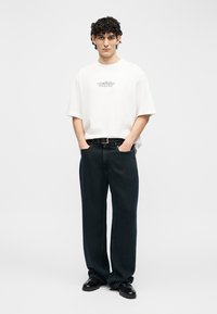 Jaunas vyras su garbanotais plaukais, vilkintis baltą oversize stiliaus marškinėlius su grafiniu printu, laisvus juodus džinsus, juodą diržą ir juodus batus, stovintis su rankomis kišenėse.