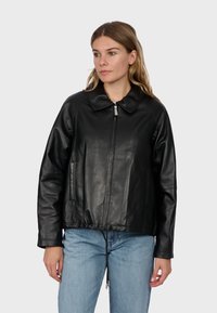 Femme aux longs cheveux portant une veste en cuir noire zippée et un jean bleu clair, debout devant un fond gris clair uni.