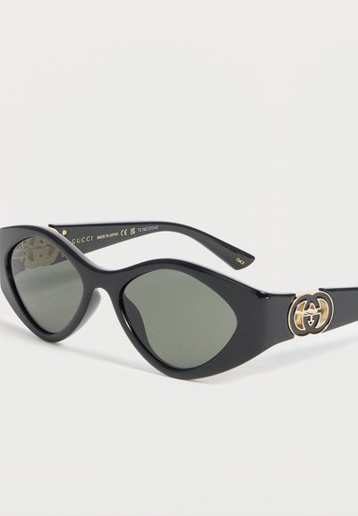 Μαύρα γυαλιά ηλίου cat-eye με χρωματικούς φακούς και χρυσό λογότυπο Gucci στο πόδι, παρουσιασμένα σε λευκό φόντο.
