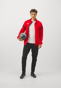 Veste de sport zippée rouge avec des accents noirs, t-shirt blanc, pantalon noir et ballon de football noir avec des détails argentés. Chaussures de sport noires.