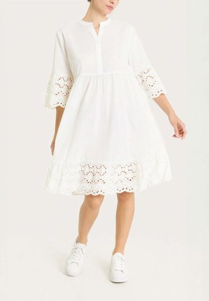 Donna che indossa un vestito bianco al ginocchio con maniche e orlo in pizzo sangallo, abbinato a sneakers bianche, in piedi su uno sfondo semplice.