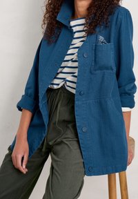 Blue denim jacket with long sleeves, two front pockets, and button closure. Worn over a navy and white striped top, paired with olive green trousers.