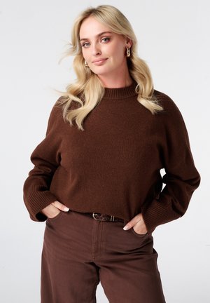 Blonde vrouw met golvend haar die gouden oorringen draagt, een bruine trui en bijpassende bruine broek met handen in de zakken.