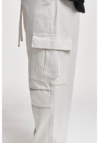 Pantalon cargo beige avec une grande poche à rabat sur le côté et des coutures visibles, porté par une personne habillée d'un haut noir.