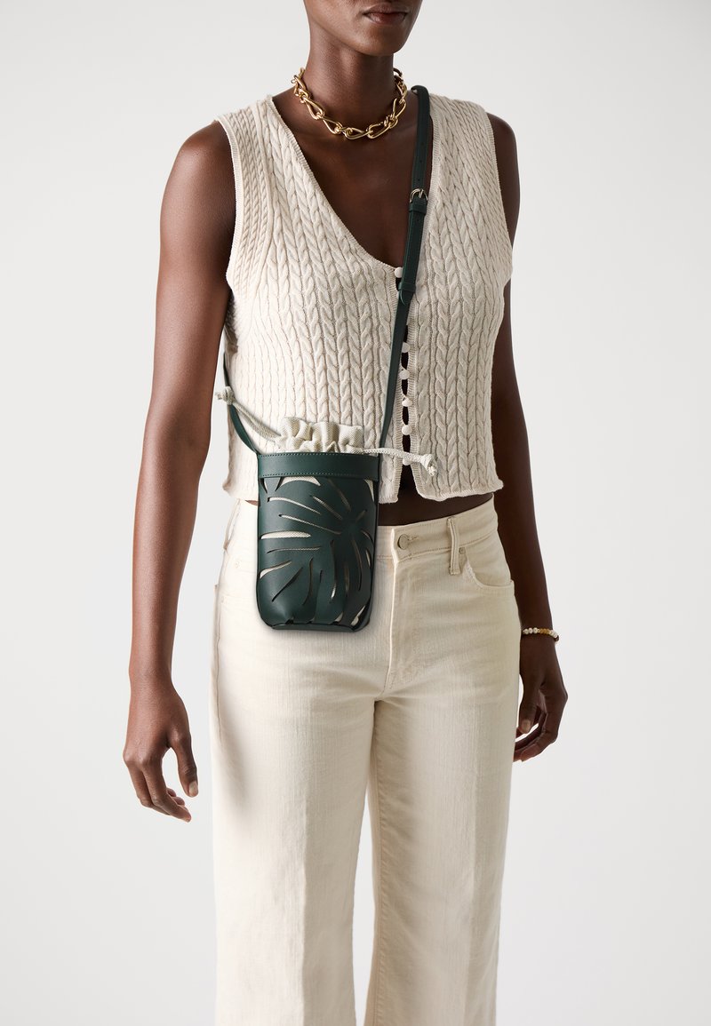 Grüne Leder-Crossbody-Tasche mit einem Schnittmuster aus Palmblättern, verstellbarem Gurt und Kordelzug oben, getragen von einer Person in hellen Hosen.
