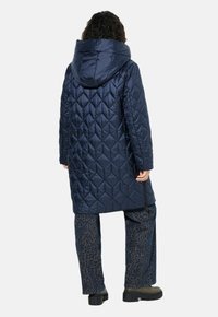 Abrigo largo acolchado en color navy con una capucha sobredimensionada, que presenta un patrón de diamantes y bolsillos laterales, usado sobre pantalones de leopardo y botas gruesas negras.