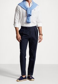 White linen shirt, navy pinstriped trousers, blue cable-knit sweater tied at neck, blue espadrilles with colorful accents.