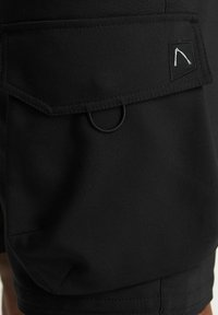 Pantaloni corti neri realizzati in tessuto liscio con una tasca a soffietto di forma rettangolare, un dettaglio ad anello e un piccolo logo ricamato in bianco.
