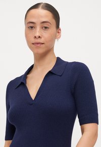 Marineblå ribbestrikket polo-stil topp med korte ermer og dyp V-hals. Stoffet har en glatt og tettsittende tekstur, med en klassisk krage design.