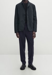 Giacca leggera nera con colletto a punta, due tasche frontali, chiusura a bottoni, abbinata a pantaloni blu navy e stivali neri. Design semplice.
