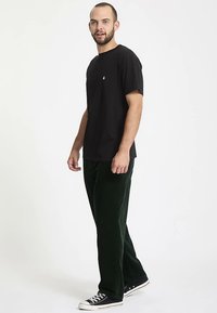 Czarny t-shirt z krótkim rękawem z małym logo, w połączeniu z ciemnozielonymi spodniami sztruksowymi i czarnymi sneakersami, stojący na neutralnym tle.