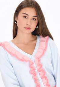 Ljusblå cardigan med rosa volangdetaljer längs halslinjen och ner längs framsidan, med ljusa knappar och en mjuk textur.