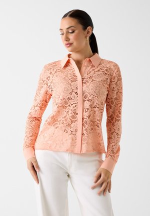 Kobieta w brzoskwiniowej koronkowej bluzce z długim rękawem, zapinanej na guziki z kołnierzykiem i białych spodniach, stojąca na jasnym tle.