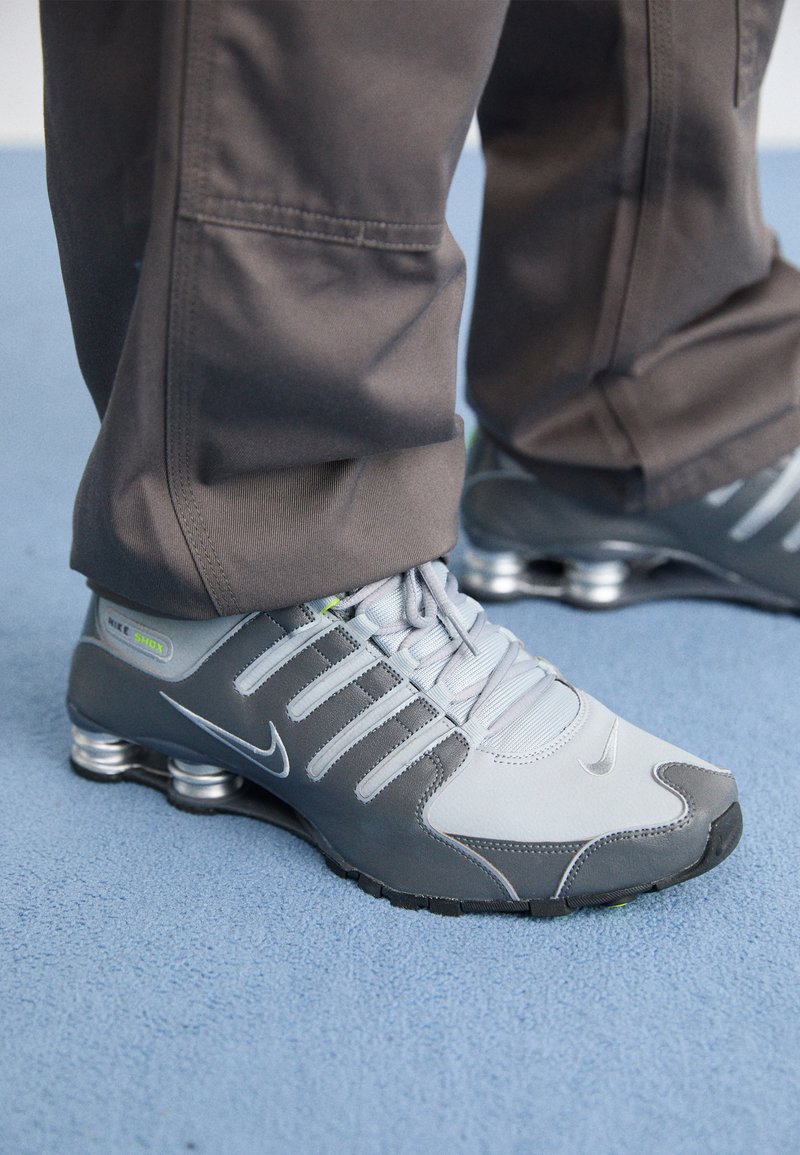 Graue und silberne Nike Shox Sneakers zeichnen sich durch ein schlankes Design aus, mit Netz- und Synthetikmaterialien, einer abgerundeten Zehenpartie und einem gepolsterten Schuhkragen.
