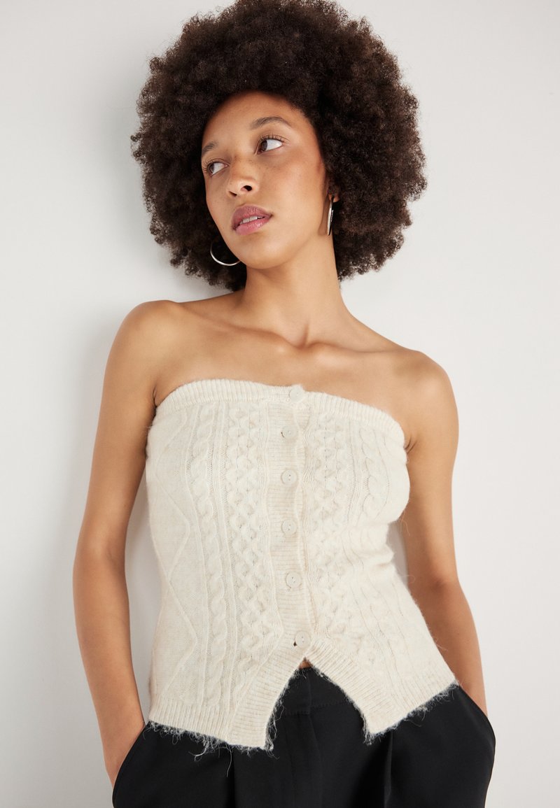 Top strapless em malha canelada creme com frente de botões, padrão texturado e bainha desfalcada. Usado com calças escuras, tecido leve e design ajustado.