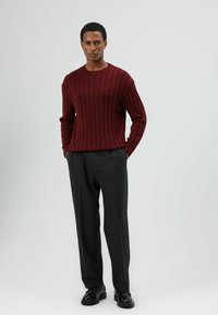 Bordeauxfarbener, gerippter Pullover mit rundem Ausschnitt, kombiniert mit dunkelgrauen, maßgeschneiderten Hosen und schwarzen Schuhen, präsentiert vor hellem Hintergrund.