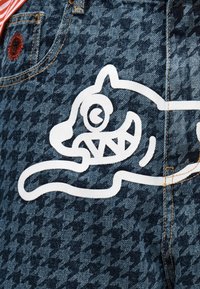 Ύφασμα ντένιμ με σκούρο μοτίβο κουκουβάγιας, που διαθέτει λευκό καρτούν γάτα με ένα μάτι και αιχμηρά δόντια κοντά σε μια μικρή τσέπη.