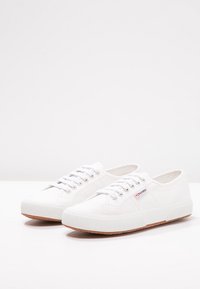 Un par de zapatillas de lona blancas bajas con cordones blancos y suelas de goma en una superficie blanca.