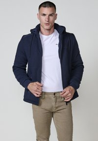 Felpa con zip della marina, con cappuccio, tessuto texture, con lacci neri, t-shirt bianca a maniche corte sotto, abbinata a pantaloni marrone chiaro.