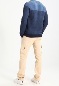 Pull en maille texturée bleu marine et bleu clair avec un ourlet côtelé, porté avec un pantalon cargo beige avec des poches latérales et des baskets blanches.