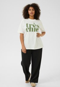 T-shirt blanc avec texte vert "très chic", coupe décontractée ; associé à un pantalon noir à jambes larges et des baskets beiges.